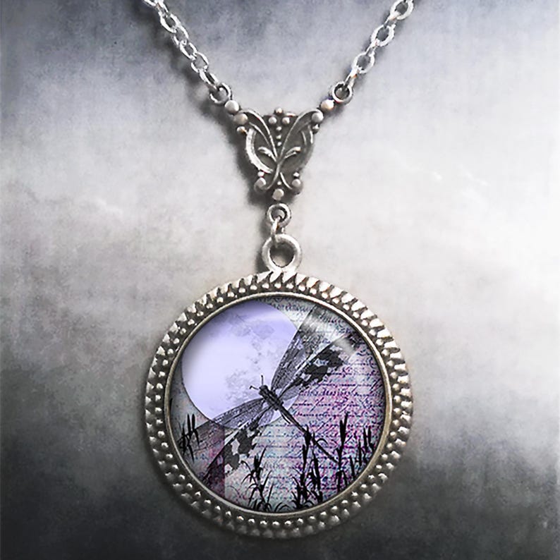 May include: Silver-toned necklace with a round pendant featuring a dragonfly design. The pendant has a lavender moon and a dragonfly silhouette over a background of script and tall grass. The necklace has a decorative bail and chain.