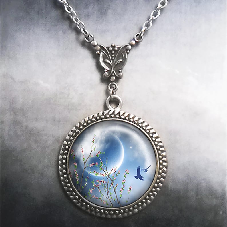 May include: A silver necklace with a round pendant featuring a crescent moon, a blooming tree branch, and a bird flying in a blue sky.