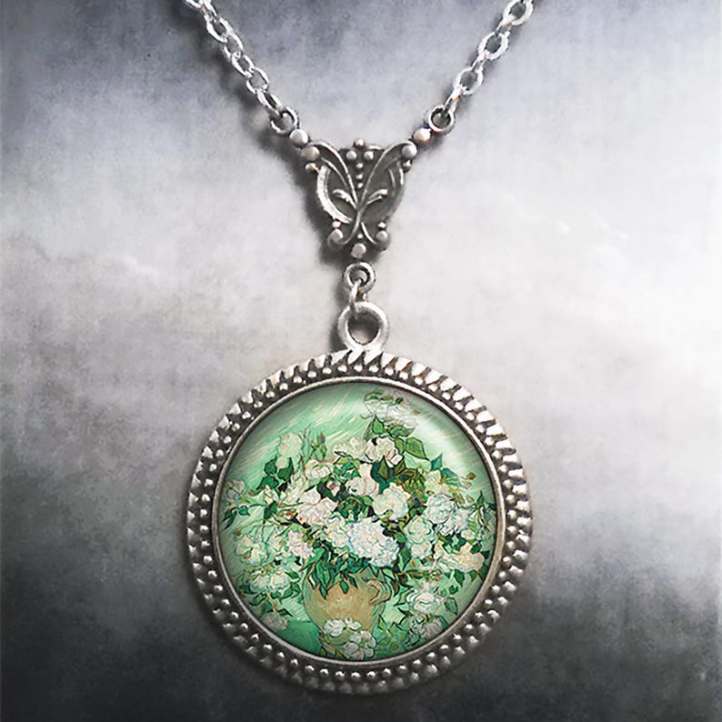 May include: A silver necklace with a round pendant featuring a painting of white flowers in a vase. The pendant has a silver frame with a beaded edge.