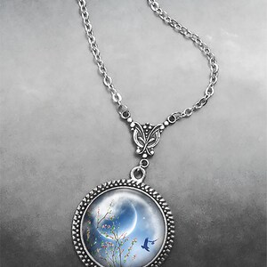 May include: A silver necklace with a round pendant featuring a blue crescent moon, a blooming tree with pink flowers, and a blue bird flying in front of the moon. The pendant is surrounded by a silver beaded frame.