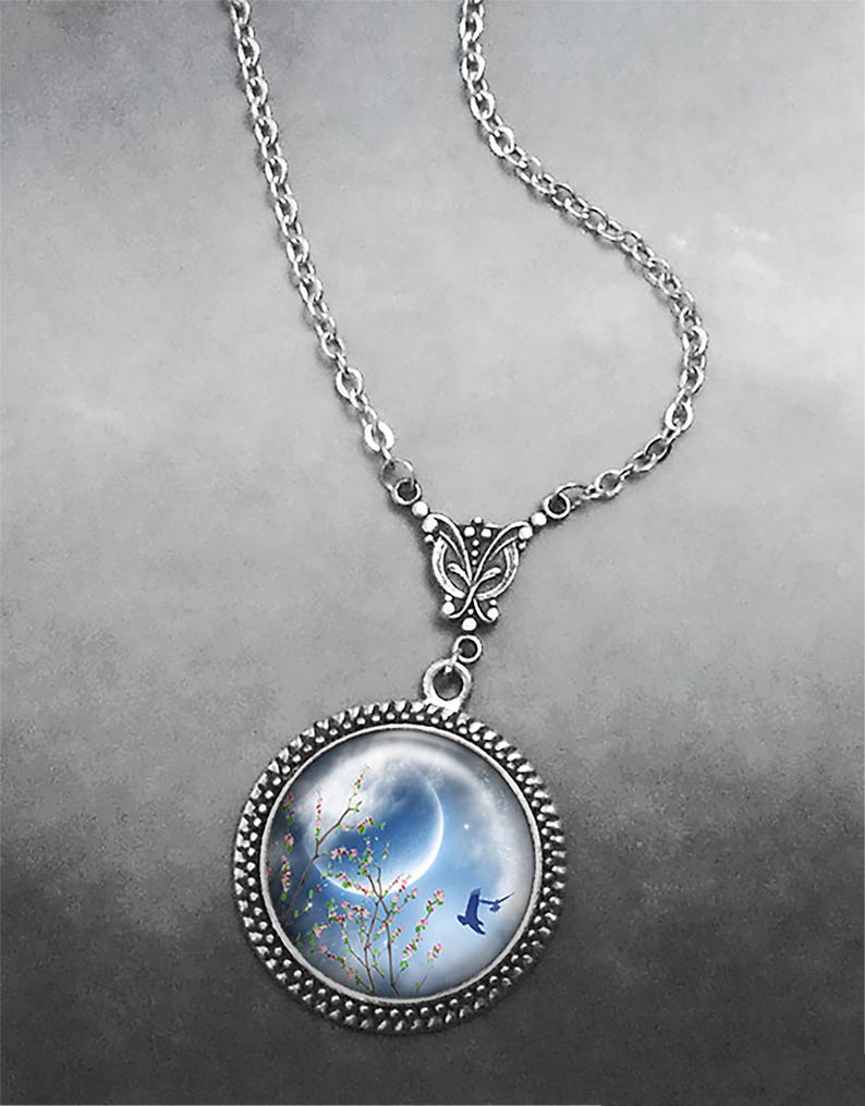 May include: A silver necklace with a round pendant featuring a blue crescent moon, a blooming tree with pink flowers, and a blue bird flying in front of the moon. The pendant is surrounded by a silver beaded frame.