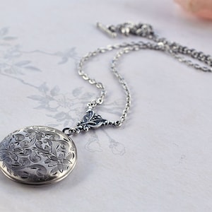 May include: A silver locket necklace with a delicate chain and a floral design on the front. The locket is round and has a detailed engraved pattern.