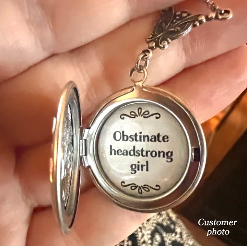 May include: Silver locket necklace with a floral design on the chain. The locket is open and the inside reads "Obstinate headstrong girl".