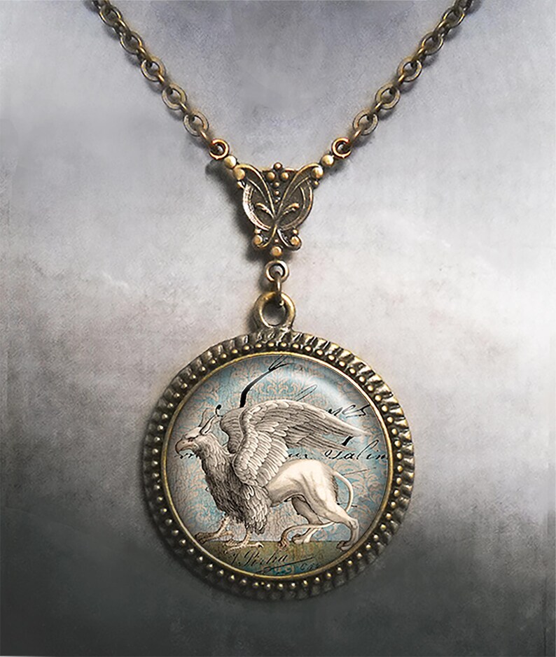 May include: A bronze necklace with a round pendant featuring a vintage illustration of a griffin. The pendant has a glass cover and a decorative border. The necklace has a butterfly-shaped clasp.