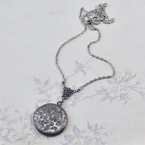 May include: A silver locket necklace with a delicate chain and a floral design. The locket is round and has a detailed engraved pattern.