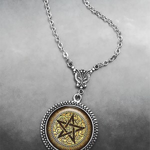 May include: A silver-toned necklace featuring a circular pendant with a textured rope-style border. The center of the pendant displays a dark brown pentagram symbol set against a gold-colored background with an intricate, swirling filigree pattern.
