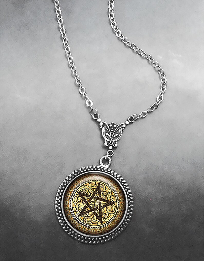 May include: A silver-toned necklace featuring a circular pendant with a textured rope-style border. The center of the pendant displays a dark brown pentagram symbol set against a gold-colored background with an intricate, swirling filigree pattern.