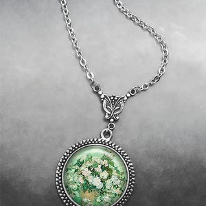May include: A silver chain necklace with a round pendant featuring a painting of white flowers in a vase. The pendant has a silver frame with a beaded edge.