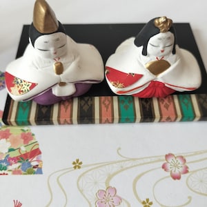 May include: Two ceramic figurines representing traditional Japanese dolls, one wearing a gold headpiece and purple base, the other with a gold hair ornament and red base. Both dolls feature white robes with red and gold patterned sleeves.