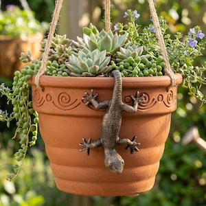 May include: A terracotta hanging planter featuring a bronze-colored lizard sculpture climbing down the side. The pot has a decorative embossed swirl pattern near the rim and is filled with various green succulents and small purple flowers.