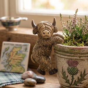 May include: A ceramic Highland cow figurine with a shaggy brown coat, perched on the edge of a weathered pot. The pot features a thistle design and holds a small plant with purple flowers. A small tile with a map of Scotland is also visible.