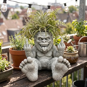 May include: A gray concrete gorilla-shaped planter with a plant growing from its head. The gorilla is seated, sticking out its tongue, and giving the middle finger with both hands. The planter is on a wooden surface, surrounded by other potted plants.