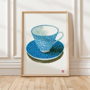 May include: A framed art print featuring a white teacup and saucer decorated with a blue leaf pattern. The teacup has a light blue interior and a handle on the right side. A small red square stamp is visible in the bottom right corner of the print.