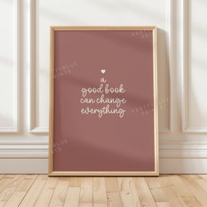 May include: A rectangular wall art print in a light wooden frame featuring a dusty rose background. The text "a good book can change everything" is written in white cursive script, centered above a small white heart icon on the print.