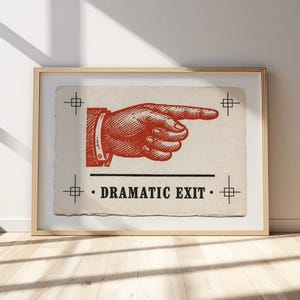May include: A framed print on off-white paper featuring a red ink illustration of a hand pointing to the right. The design includes four small crosshair symbols in the corners and the black text "DRAMATIC EXIT" centered below the hand.