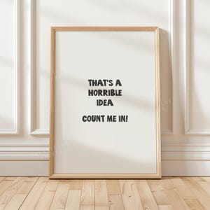 May include: A rectangular wall art print in a light wood frame featuring a white background with black text that reads: THAT'S A HORRIBLE IDEA COUNT ME IN! The print is centred and displays bold, sans-serif lettering in a clean, modern layout.