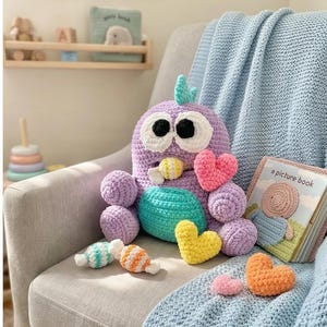 Puede incluir: Un peluche de búho de ganchillo hecho a mano en tonos morado, turquesa y blanco, con grandes ojos negros y un pico amarillo. El búho está rodeado de corazones y caramelos de ganchillo, y un libro infantil.