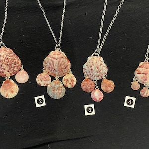 May include: Four seashell necklaces with silver chains. Each necklace features a large, textured shell pendant with smaller shell accents in shades of pink, orange, and white. The necklaces are displayed on a black surface.