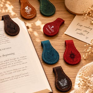 May include: A collection of teardrop-shaped leather magnetic bookmarks in various colors including brown, red, blue, and green. Each bookmark features a unique white engraved design, such as a cross, a flower, or a bird, with some including scripture references.