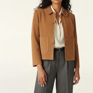 Könnte beinhalten: Hellbraune Jacke mit rundem Kragen und zwei Vordertaschen. Die Jacke hat einen Knopfverschluss und wird mit grauer Hose und weißer Bluse kombiniert. Das Outfit wird durch einen schwarzen Gürtel ergänzt.