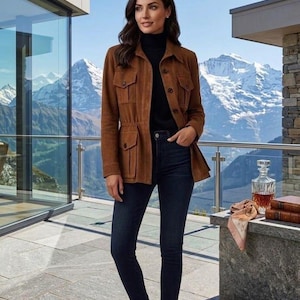 Könnte beinhalten: Eine Frau trägt eine braune Wildlederjacke mit mehreren Taschen, einen schwarzen Rollkragenpullover und dunkelblaue Jeans. Die Jacke hat einen Kordelzug in der Taille und Knöpfe. Sie steht auf einer Terrasse mit Bergpanorama.