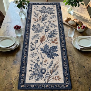 May include: A rectangular table runner featuring a cream-coloured centre panel with a detailed botanical print of blue and brown oak leaves, acorns, and wheat stalks. The runner is framed by a dark blue border with a small floral pattern.