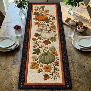 Könnte beinhalten: Ein rechteckiger, gesteppter Tischläufer mit einem cremefarbenen Mittelteil, der mit einem gestickten Muster aus orangefarbenen, weißen und grünen Kürbissen, Herbstblättern und Blumen verziert ist. Er hat einen breiten orangefarbenen Rand.