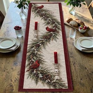 May include: A rectangular quilted table runner featuring a cream-colored center with a printed design of evergreen boughs, pinecones, red berries, three red cardinals, and two red pillar candles in white distressed candle holders, bordered in red.