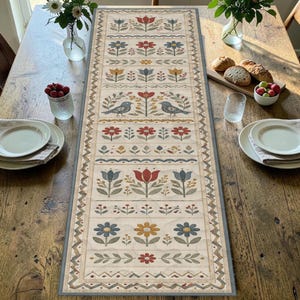 May include: A rectangular table runner featuring a cream-colored background with repeating horizontal bands of folk-art style floral motifs, leaves, and two birds facing each other, all bordered by a gray geometric pattern trim.