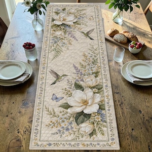 May include: A rectangular table runner featuring a cream-colored background with a repeating honeycomb texture. The design displays large white magnolia blossoms, green foliage, small blue flowers, two hummingbirds in flight, and a blue butterfly.