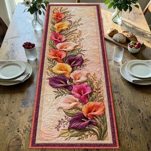 Op de afbeelding: Een rechthoekige, doorgestikte tafelloper met een centraal bloemmotief van veelkleurige calla-lelies in tinten oranje, geel, roze en paars. De achtergrond is van crèmekleurige stof met ingewikkelde, wervelende stikpatronen.