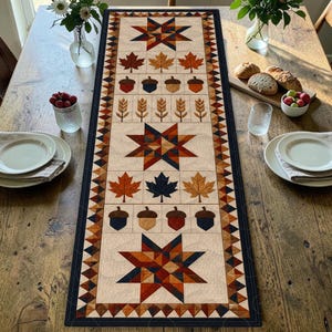 May include: A rectangular quilted table runner featuring a cream background with a repeating pattern of stars, maple leaves, acorns, and wheat stalks in autumn colours of orange, brown, navy blue, and gold, bordered by a dark navy blue triangle trim.