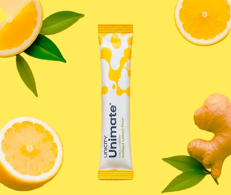 K&ouml;nnte beinhalten: Ein wei&szlig;es Unimate-P&auml;ckchen mit gelben Mustern und dem Text "Unicity Unimate Nootropic Lemon Flavor" auf gelbem Hintergrund. Zitronenscheiben, gr&uuml;ne Bl&auml;tter und ein St&uuml;ck Ingwer sind ebenfalls vorhanden.