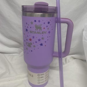 May include: A lavender Stanley 1.18 litre tumbler with a handle and a matching lid. The tumbler features a constellation design with stars and butterflies, and the Stanley logo. A clear straw rests against the side.