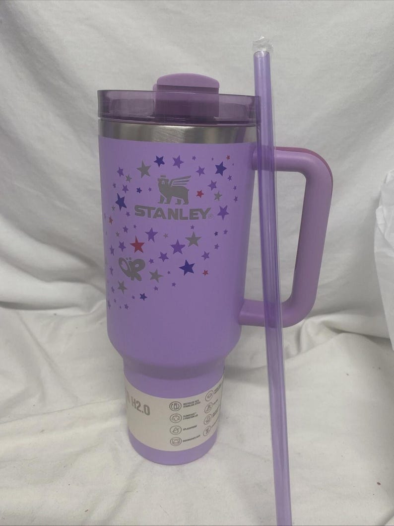 May include: A lavender Stanley 1.18 litre tumbler with a handle and a matching lid. The tumbler features a constellation design with stars and butterflies, and the Stanley logo. A clear straw rests against the side.
