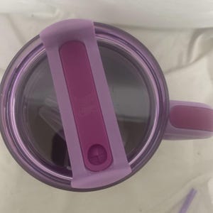 May include: A purple travel mug with a matching lid and handle. The lid has a flip-top closure in a darker shade of purple. The mug appears to be made of clear plastic, allowing the contents to be visible. The handle is a solid, curved design.