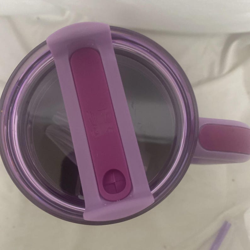 May include: A purple travel mug with a matching lid and handle. The lid has a flip-top closure in a darker shade of purple. The mug appears to be made of clear plastic, allowing the contents to be visible. The handle is a solid, curved design.