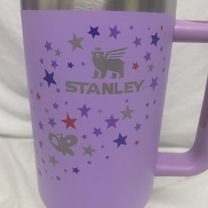 May include: A lilac Stanley mug with a silver rim and handle. The mug features a pattern of stars in shades of purple and red. The Stanley logo is visible on the front.
