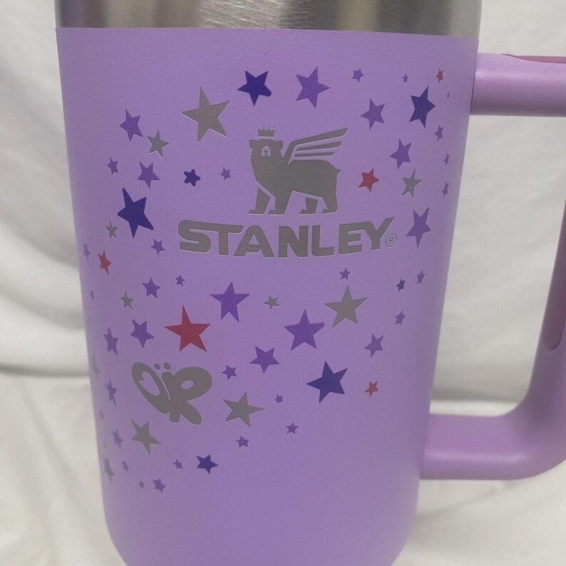 May include: A lilac Stanley mug with a silver rim and handle. The mug features a pattern of stars in shades of purple and red. The Stanley logo is visible on the front.