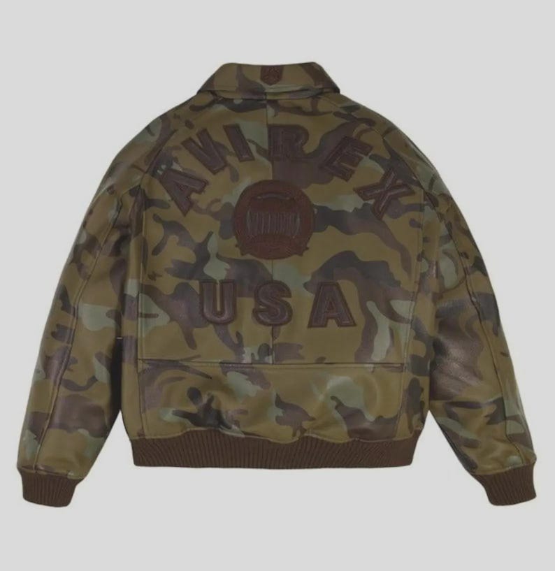 May include: A camouflage print leather bomber jacket featuring a brown collar, cuffs, and hem. The back displays the dark brown embossed text AVIREX USA in a curved arrangement above a circular emblem, all set against a green and brown pattern.