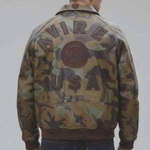 May include: A camouflage print leather jacket featuring the text AVIREX and USA in dark brown leather appliqu&eacute; on the back. The jacket includes a circular emblem in the center and a ribbed brown knit waistband and sleeve cuffs.