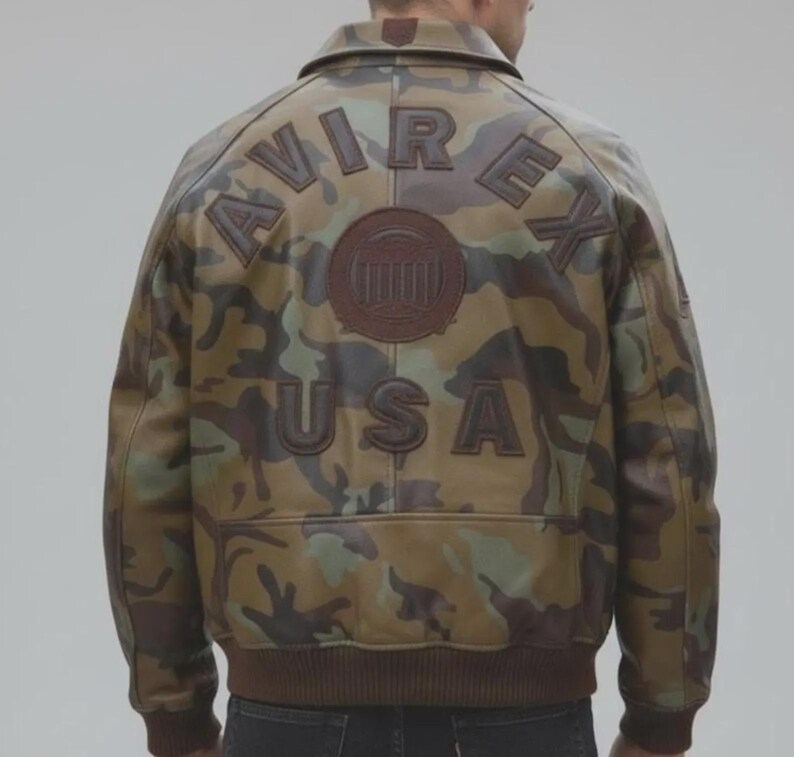 May include: A camouflage print leather jacket featuring the text AVIREX and USA in dark brown leather appliqu&eacute; on the back. The jacket includes a circular emblem in the center and a ribbed brown knit waistband and sleeve cuffs.
