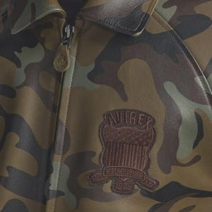 May include: A camouflage print leather jacket featuring a brown leather patch on the chest with the text AVIREX, ESTABLISHED 1975, and NY. The jacket has a full-length front zipper with a branded leather pull tab and a classic collar design.