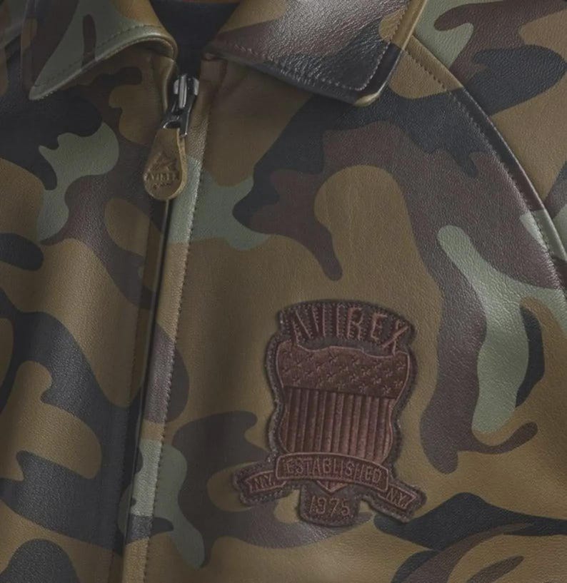 May include: A camouflage print leather jacket featuring a brown leather patch on the chest with the text AVIREX, ESTABLISHED 1975, and NY. The jacket has a full-length front zipper with a branded leather pull tab and a classic collar design.