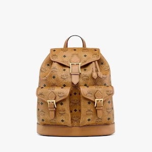 May include: A brown backpack with a repeating pattern of black diamonds and the letters "MCM". The backpack has two front pockets with gold buckles, a top handle, and a drawstring closure. The base of the backpack is a darker shade of brown.