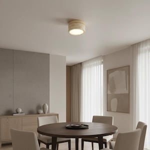 May include: A round, beige ceiling light fixture illuminates a dining room. The light is mounted on a white ceiling. A dark wood dining table is surrounded by beige chairs. The room has neutral tones and a minimalist design.