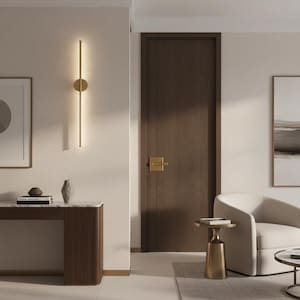 May include: A modern interior scene featuring a wall-mounted gold light fixture with a vertical bar and circular accent. A dark wood door with a gold handle is visible, along with a white armchair and a small side table.