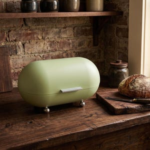 Op de afbeelding: Een saliegroene brooddoos met een afgeronde rechthoekige vorm, zilveren handvat en kleine zilveren voetjes. De brooddoos staat op een houten oppervlak, met een brood en een houten snijplank in de buurt.