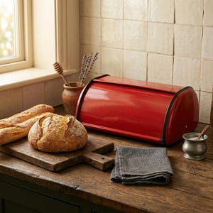 May include: A red bread bin with black trim is on a wooden surface alongside a loaf of bread and a baguette. A wooden chopping board and a grey cloth are also on the table. A small pot of salt and a honey dipper are also visible.