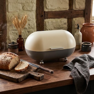 May include: A two-tone bread box in cream and gray sits on a wooden countertop. A loaf of bread, a cutting board, and a knife are in the foreground. Other kitchen items include a brown bottle, a small jar, and a ceramic pot.